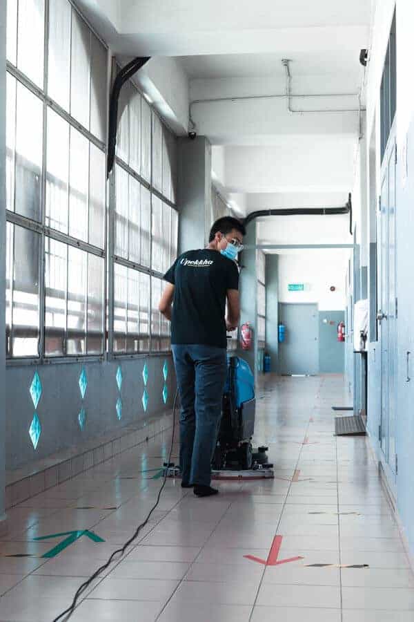 A man is pushing Upekkha's UpSweep E3040 auto scrubber machine in a brightly lit walkway on brown floor tiles.